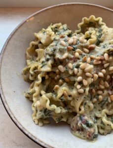 Pasta in een beige kom. In de kom zie je de groene stukjes van spinazie en bovenop pijnboompitten gestrooid.