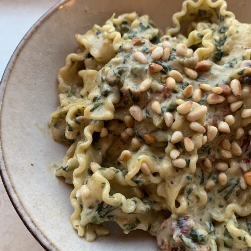 Pasta in een beige kom. In de kom zie je de groene stukjes van spinazie en bovenop pijnboompitten gestrooid.